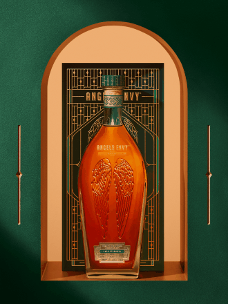 Angel’s Envy 2026 Cask Strength Bourbon bottle displayed in front of the limited-edition presentation box,  framed by an arched niche within the Cask Archive room and set in the Cask Archives room.